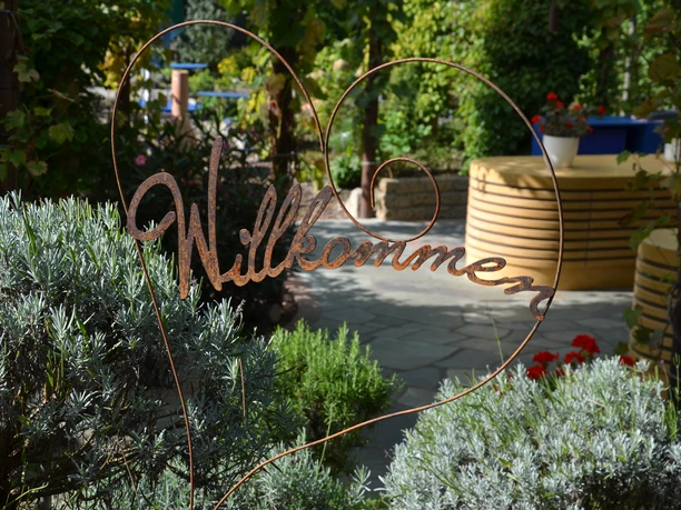 Rila erleben Ein herzförmiges Metallschild mit "Willkommen", umgeben von grünen Pflanzen in einem Garten.