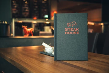 Espelkamp Steakhouse.jpg Steakhouse-Menü auf Holztisch mit stimmungsvoller Beleuchtung und eleganter Einrichtung im Hintergrund.