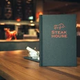 Espelkamp Steakhouse.jpg Steakhouse-Menü auf Holztisch mit stimmungsvoller Beleuchtung und eleganter Einrichtung im Hintergrund.