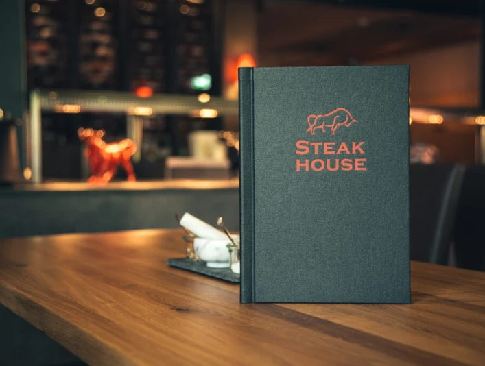 Espelkamp Steakhouse.jpg Steakhouse-Menü auf Holztisch mit stimmungsvoller Beleuchtung und eleganter Einrichtung im Hintergrund.