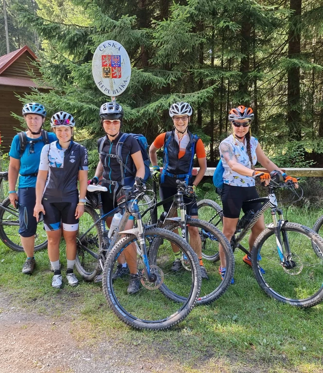 A group of cyclists pose with mountain bikes in front of a forest hut on a sunny day.