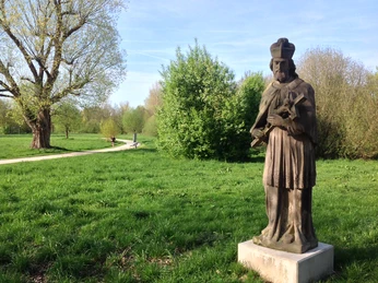 Steinfigur eines Bischofs auf grünem Rasen, umgeben von Bäumen, mit Spazierweg im Hintergrund.