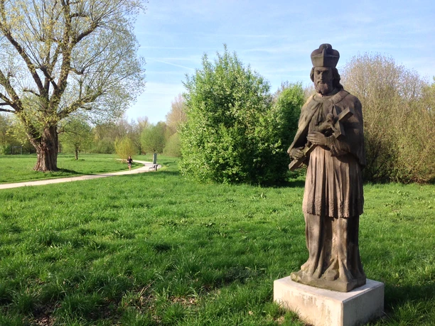 Steinfigur eines Bischofs auf grünem Rasen, umgeben von Bäumen, mit Spazierweg im Hintergrund.