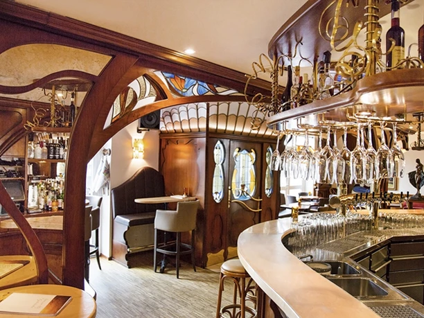 Gemütliche Bar im Jugendstil, geschwungene Holzelemente, stilvolle Hängelampen, einladende Atmosphäre.