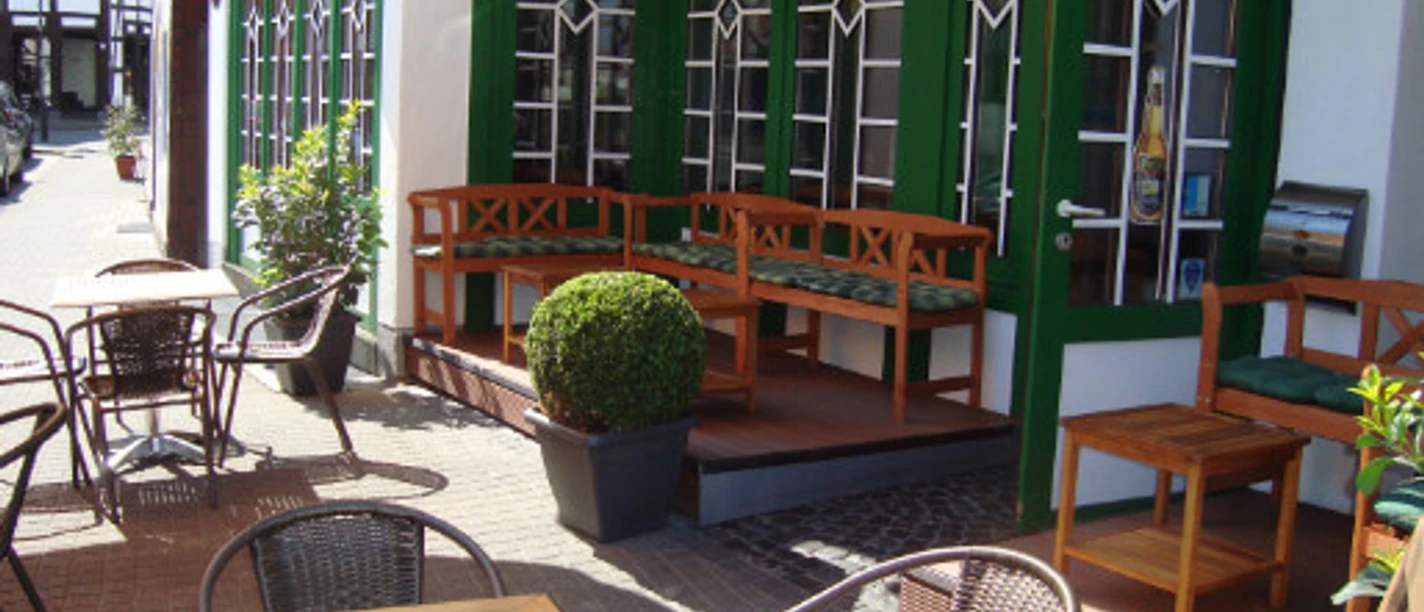 Gemütliche Außenterrasse eines Cafés mit Holztischen, Stühlen und Pflanzen vor großen Fenstern.