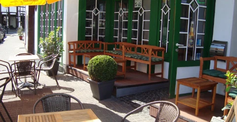 Pub.jpg Gemütliche Außenterrasse eines Cafés mit Holztischen, Stühlen und Pflanzen vor großen Fenstern.