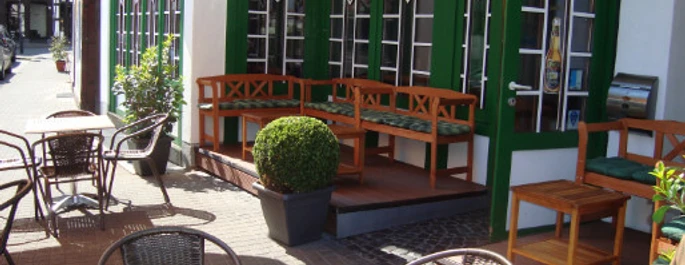 Pub.jpg Gemütliche Außenterrasse eines Cafés mit Holztischen, Stühlen und Pflanzen vor großen Fenstern.