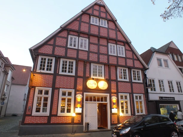 Delphi.jpg Fachwerkhaus bei Dämmerung, rot-weiße Ziegel, weiße Fensterrahmen, beleuchtete Gasthausreklame.