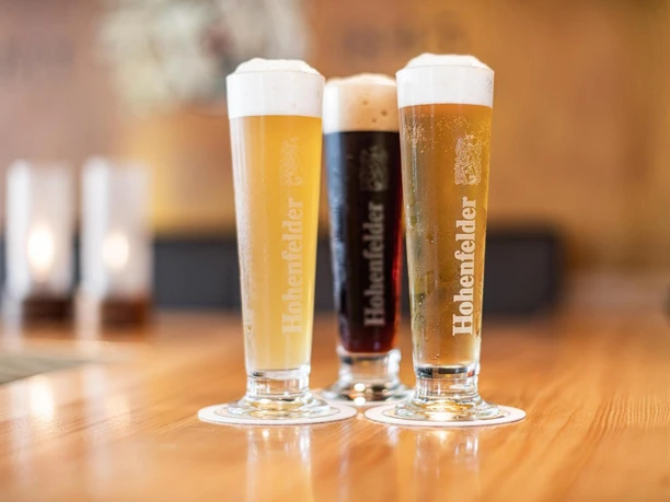 Drei gefüllte Biergläser mit markanter Schaumkrone auf einem Holztresen, im Hintergrund unscharf.