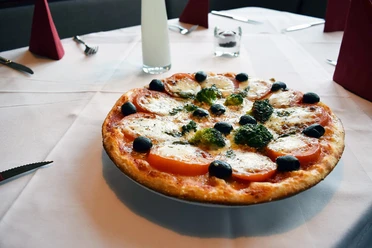 Pizza Margherita mit Tomaten, Mozzarella, Oliven und Kräutern auf weiß gedecktem Tisch angerichtet.