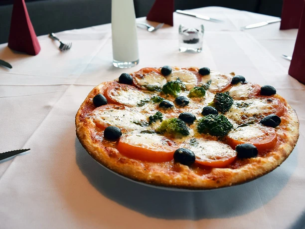 Pizza Margherita mit Tomaten, Mozzarella, Oliven und Kräutern auf weiß gedecktem Tisch angerichtet.
