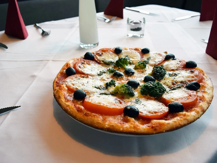 filippo_1.jpg Pizza Margherita mit Tomaten, Mozzarella, Oliven und Kräutern auf weiß gedecktem Tisch angerichtet.