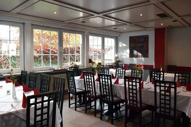 Ein helles Restaurant mit gedeckten Tischen, weißen Tischdecken und roten Servietten dekoriert.