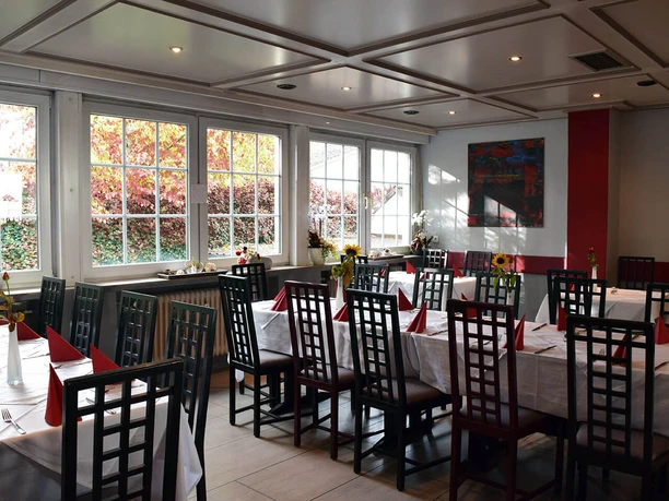 Ein helles Restaurant mit gedeckten Tischen, weißen Tischdecken und roten Servietten dekoriert.