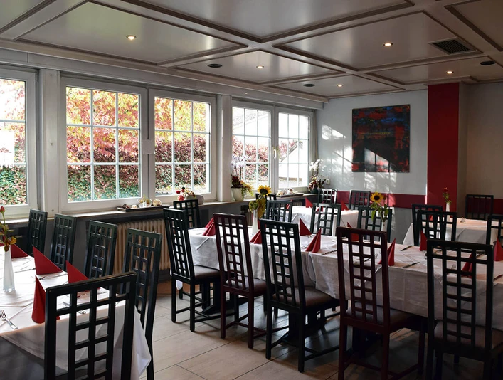 Ein helles Restaurant mit gedeckten Tischen, weißen Tischdecken und roten Servietten dekoriert.