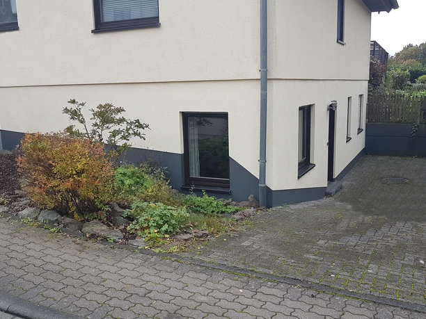 Aussenansicht.jpg Einfamilienhaus mit heller Fassade, Kellerfenstern, steiler gepflasterter Einfahrt und gepflegter Bepflanzung.