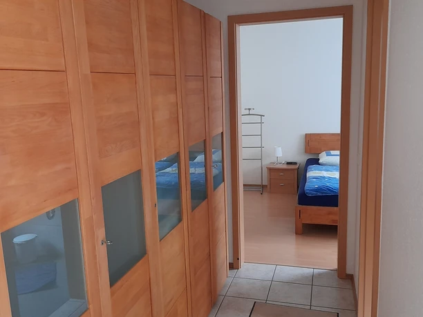 Flur.jpg Flur mit Holzschränken, führt zu einem Schlafzimmer mit Holzmöbeln und blau-weiß gestreifter Bettwäsche.