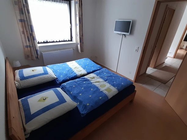 Schlafzimmer.jpg Helles Schlafzimmer mit Doppelbett und blauen Bettdecken, Fenster mit Vorhängen und TV an der Wand.