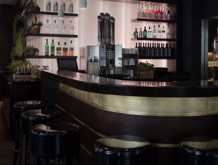 Königs_3.png Elegante Bar mit dunklem Tresen, umgeben von schwarzen Barhockern, im sanften Licht beleuchtet.