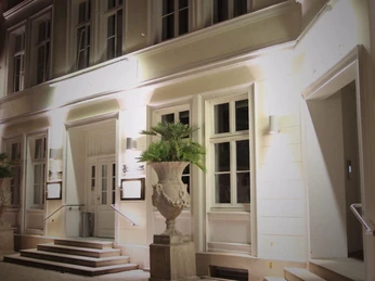 Königs_2.png Elegante Fassade eines historischen Gebäudes bei Nacht mit warm beleuchteten Fenstern und Palmen.