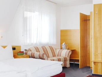 Gemütliches, helles Hotelzimmer mit Doppelbett, gestreiftem Sofa, Schreibtisch und offenem Waschbereich.