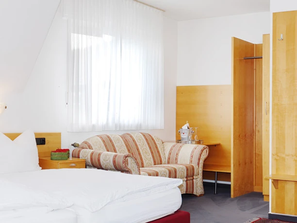 Gemütliches, helles Hotelzimmer mit Doppelbett, gestreiftem Sofa, Schreibtisch und offenem Waschbereich.