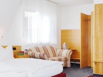 Pöppelbaum_3.jpg Gemütliches, helles Hotelzimmer mit Doppelbett, gestreiftem Sofa, Schreibtisch und offenem Waschbereich.