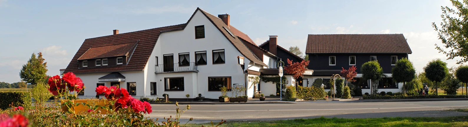 Pöppelbaum_1.jpg Großer, traditioneller Gasthof mit weißer Fassade und rotem Ziegeldach, umgeben von blühenden Rosen.