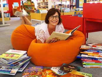 Bibliothek_Menschen.jpg Eine Frau liegt entspannt auf einem orangefarbenen Sitzsack in einer Bibliothek und liest ein Buch.