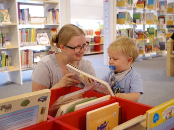 Eine Mutter zeigt ihrem kleinen Sohn ein Bilderbuch in einer hellen, freundlichen Bibliothek.