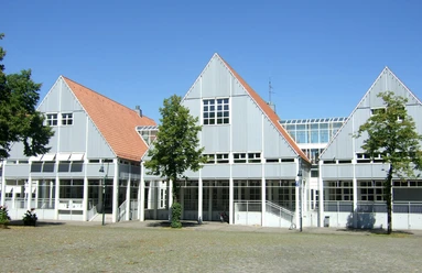 Bibliothek_Stadthaus.jpg Grau-weißes Gebäude mit Giebeldächern und großen Fensterfronten vor blauem Himmel, umgeben von Bäumen.