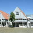 Bibliothek_Stadthaus.jpg Grau-weißes Gebäude mit Giebeldächern und großen Fensterfronten vor blauem Himmel, umgeben von Bäumen.