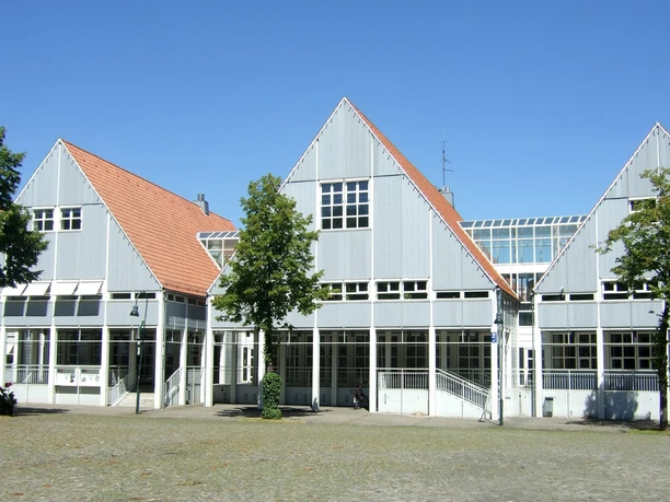 Bibliothek_Stadthaus.jpg Grau-weißes Gebäude mit Giebeldächern und großen Fensterfronten vor blauem Himmel, umgeben von Bäumen.