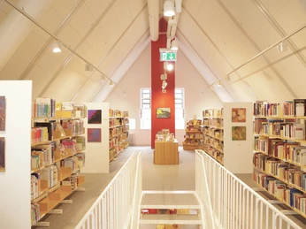 Heller Bibliotheksraum mit geneigtem Dach, Bücherregalen, weißen Wänden und einem roten Wandakzent.