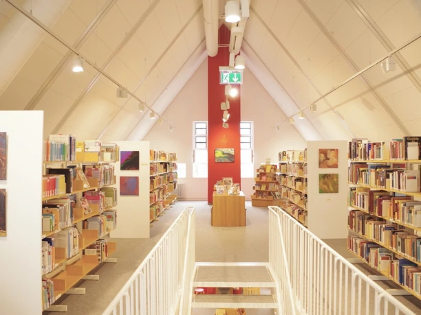 Bibliothek_Stadthaus_innen.jpg Heller Bibliotheksraum mit geneigtem Dach, Bücherregalen, weißen Wänden und einem roten Wandakzent.