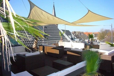 Dachterrasse mit modernen Rattanmöbeln, Sonnensegeln und Blick auf Kirchturm und umliegende Dächer.