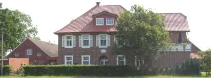 Backsteinhaus mit rotem Dach in ländlicher Umgebung, üppiges Grün und blauer Himmel im Hintergrund.