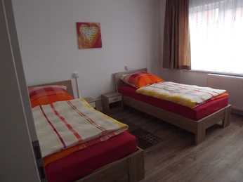 Zwei Einzelbetten mit bunten Bettdecken in einem schlicht eingerichteten, hellen Zimmer mit Holzfußboden.