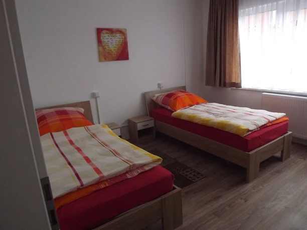 Zwei Einzelbetten mit bunten Bettdecken in einem schlicht eingerichteten, hellen Zimmer mit Holzfußboden.