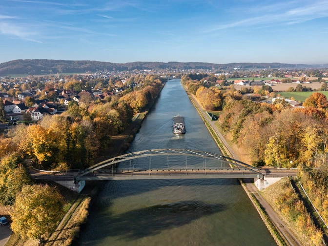 Brücke über einen herbstlichen Fluss mit Frachtschiff, umgeben von buntem Laub.Bridge over an autumnal river with a cargo ship, surrounded by colorful foliage.Bro over en efterårsagtig flod med et fragtskib, omgivet af farverigt løv.Brug over een herfstige rivier met een vrachtschip, omringd door kleurrijk gebladerte.
