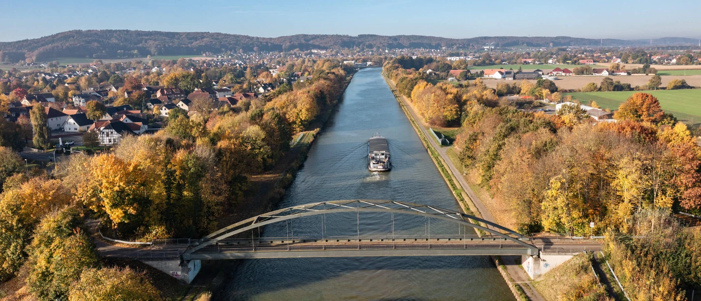 Panoramablick auf den Mittellandkanal bei Bad Essen Brug over een herfstige rivier met een vrachtschip, omringd door kleurrijk gebladerte.