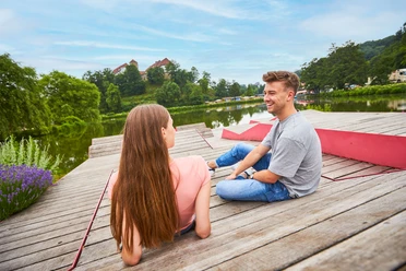 Een man en een vrouw zitten ontspannen op een houten steiger bij het meer, groene bomen op de achtergrond.