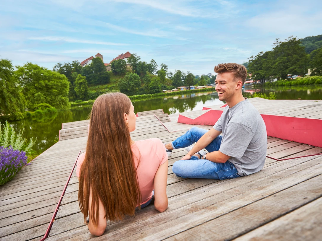 Relaxen am Charlottensee in Bad Iburg Ein Mann und eine Frau sitzen entspannt auf einem Holzsteg am See, im Hintergrund grüne Bäume.A man and a woman sit relaxed on a wooden jetty by the lake, green trees in the background.En mand og en kvinde sidder afslappet på en træbrygge ved søen med grønne træer i baggrunden.Een man en een vrouw zitten ontspannen op een houten steiger bij het meer, groene bomen op de achtergrond.
