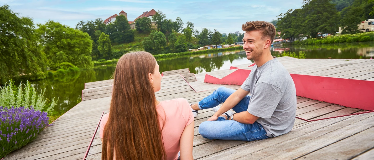 Relaxen am Charlottensee in Bad Iburg A man and a woman sit relaxed on a wooden jetty by the lake, green trees in the background.