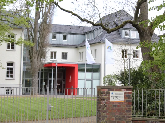 Jugendherberge Petershagen Jugendherberge Petershagen mit modernem Eingang, zwei Flaggen und grünem Rasen, umgeben von Bäumen.