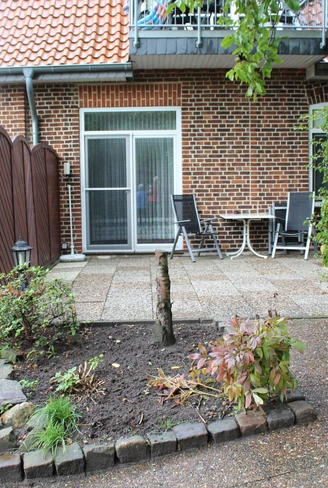 Ziegelhaus mit heller Terrasse, umgeben von Pflanzen und Gartenmöbeln, bei sonnigem Wetter.