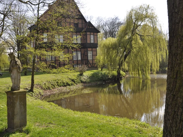 Fachwerkhaus am ruhigen Fluss, umgeben von üppigen Weiden, daneben eine Statue im grünen Park.