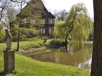 Haus Aussell Fachwerkhaus am ruhigen Fluss, umgeben von üppigen Weiden, daneben eine Statue im grünen Park.