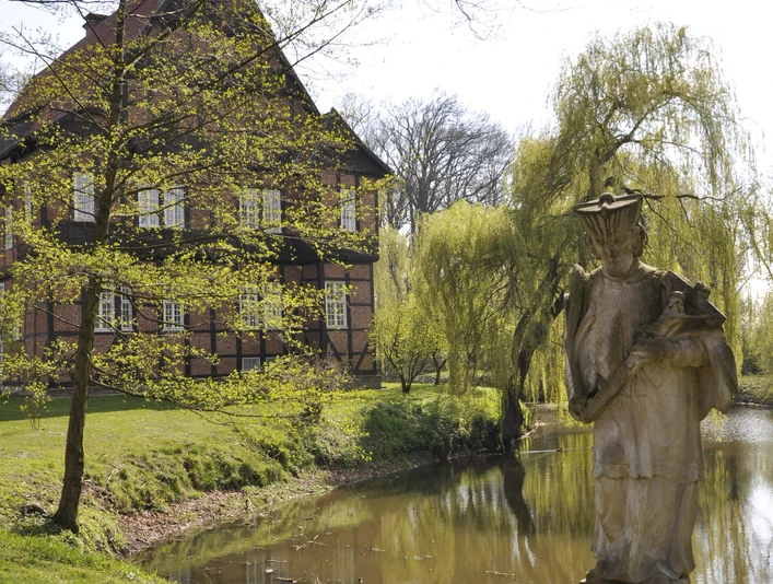 Haus Aussel Historisches Fachwerkhaus am Teich, flankiert von einer Steinfigur, umgeben von frühlinghafter Natur.