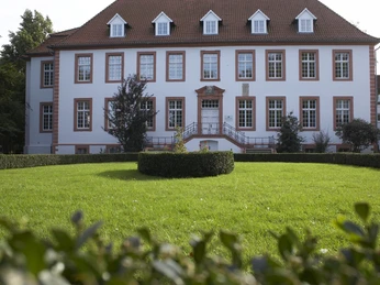 Ein historisches, zweigeschossiges Gebäude mit roten Dachziegeln und weißen Fensterrahmen, umgeben von gepflegten Gärten.